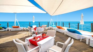 Eine Terrasse mit weißen Korbmöbeln, roten und blauen Kissen sowie einem Esstisch mit Meerblick.