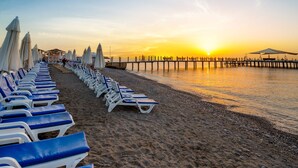 Ein Strand mit Reihen von blauen und weißen Liegestühlen und Sonnenschirmen, eine hölzerne Mole, die sich ins Wasser erstreckt, und eine untergehende Sonne im Hintergrund.