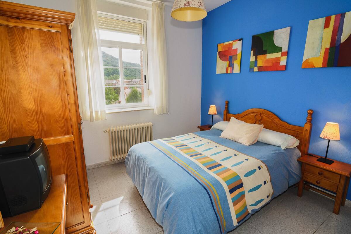 Une chambre avec un lit en bois, un radiateur, une fenêtre avec des rideaux et des tableaux abstraits accrochés au mur.