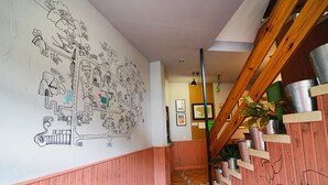 A hallway with a wooden staircase, a mural on the wall, and potted plants. - Albergue Cueva de Valporquero (Vegacervera)