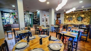 A restaurant with multiple dining tables and chairs, a kitchen area in the background, and a stone wall with decorative plates. - Albergue Cueva de Valporquero (Vegacervera)