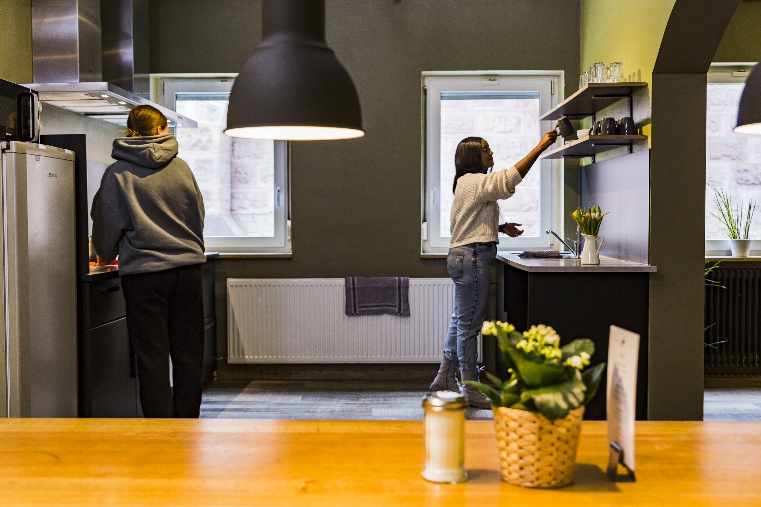 Une cuisine moderne où deux personnes travaillent, avec un plan de travail en bois et une plante en pot.