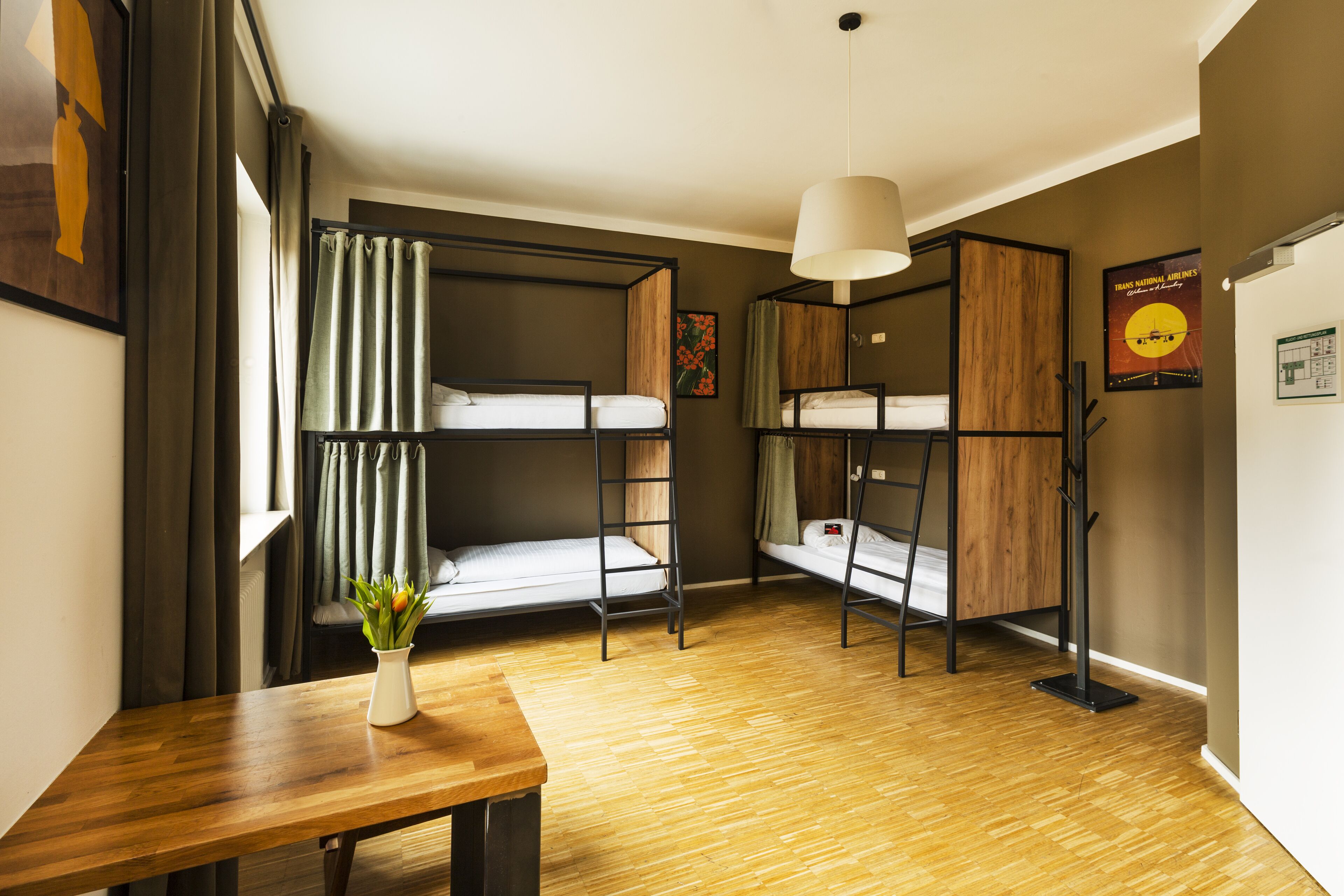 A dormitory room with bunk beds, a wooden table, a hanging light fixture, and a poster on the wall.