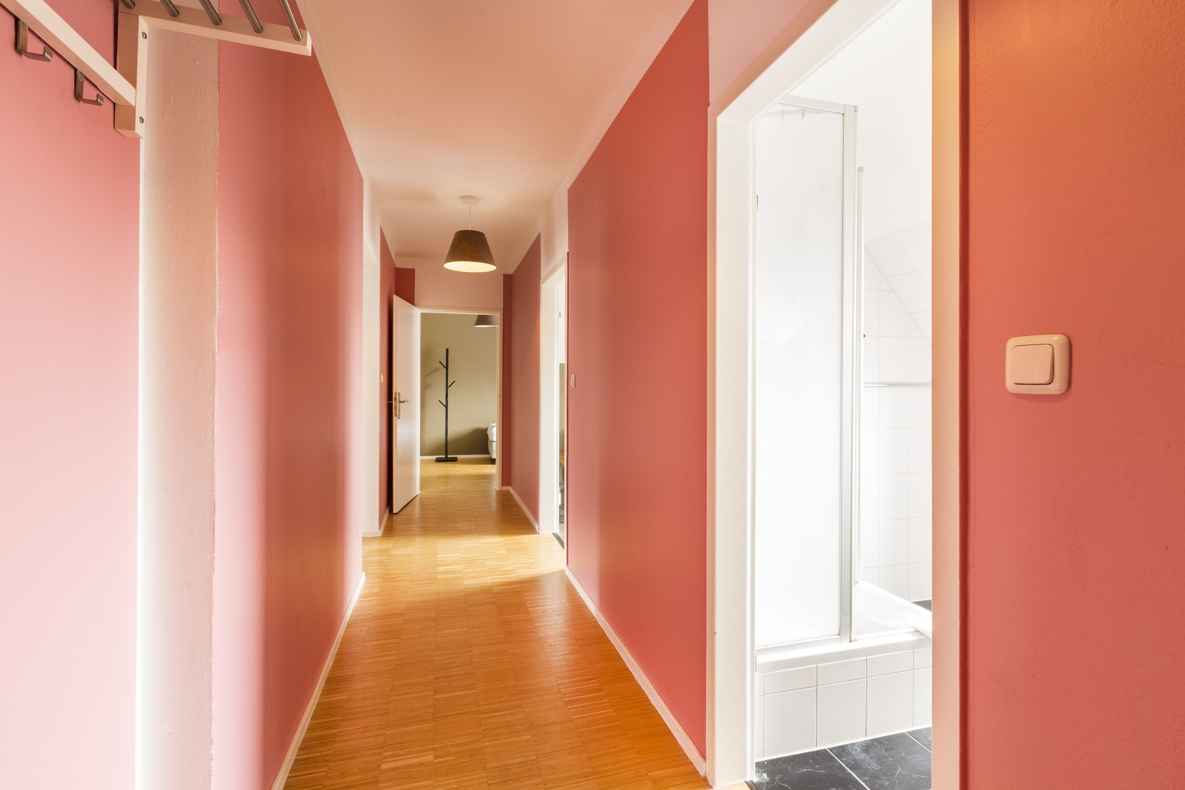 A hallway with pink walls and wooden flooring leading to a bathroom with a glass shower.