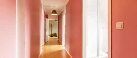 A hallway with pink walls and wooden flooring leading to a bathroom with a glass shower.