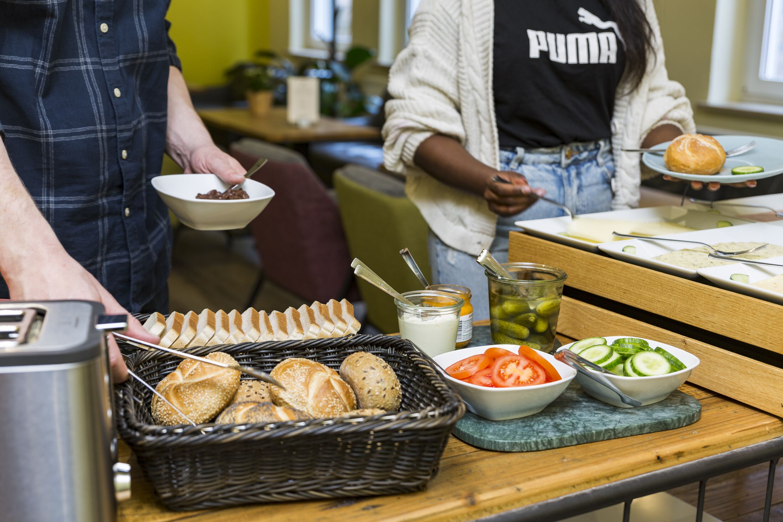 A buffet setup with various food items including bread, tomatoes, cucumbers, and a muffin.