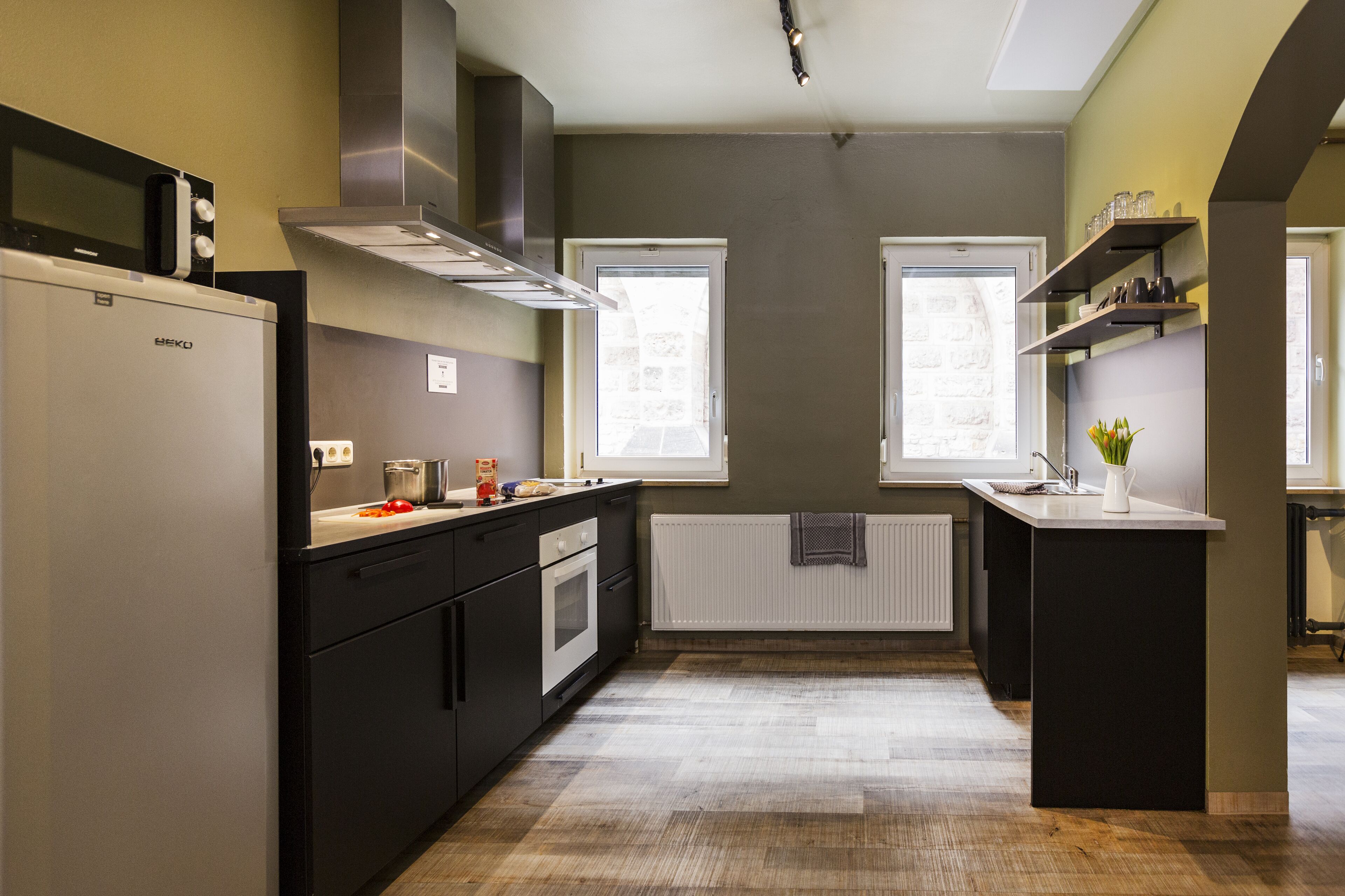 A modern kitchen with a built-in refrigerator, oven, and microwave. There are two windows providing natural light, and a small island with a vase of flowers.