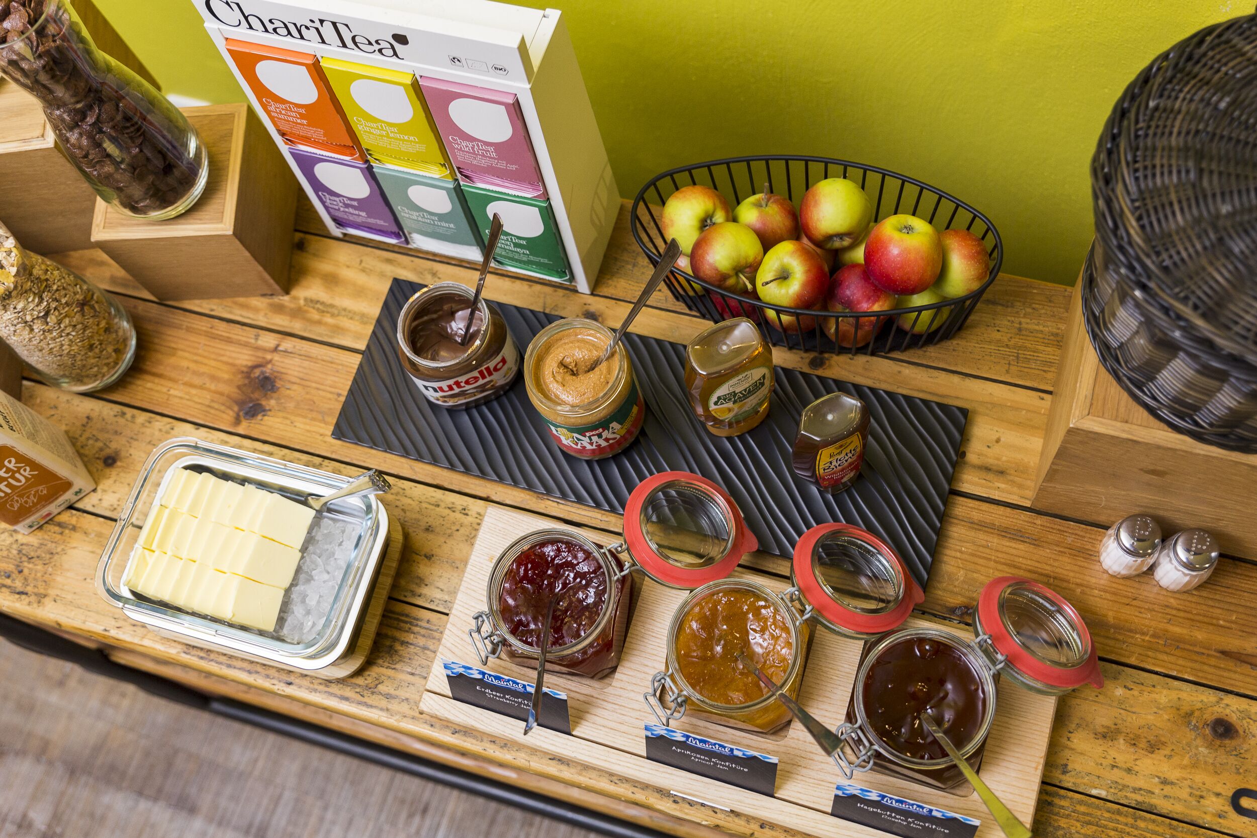 A wooden table with jars of spreads, a bowl of apples, and a box of Charitea products.