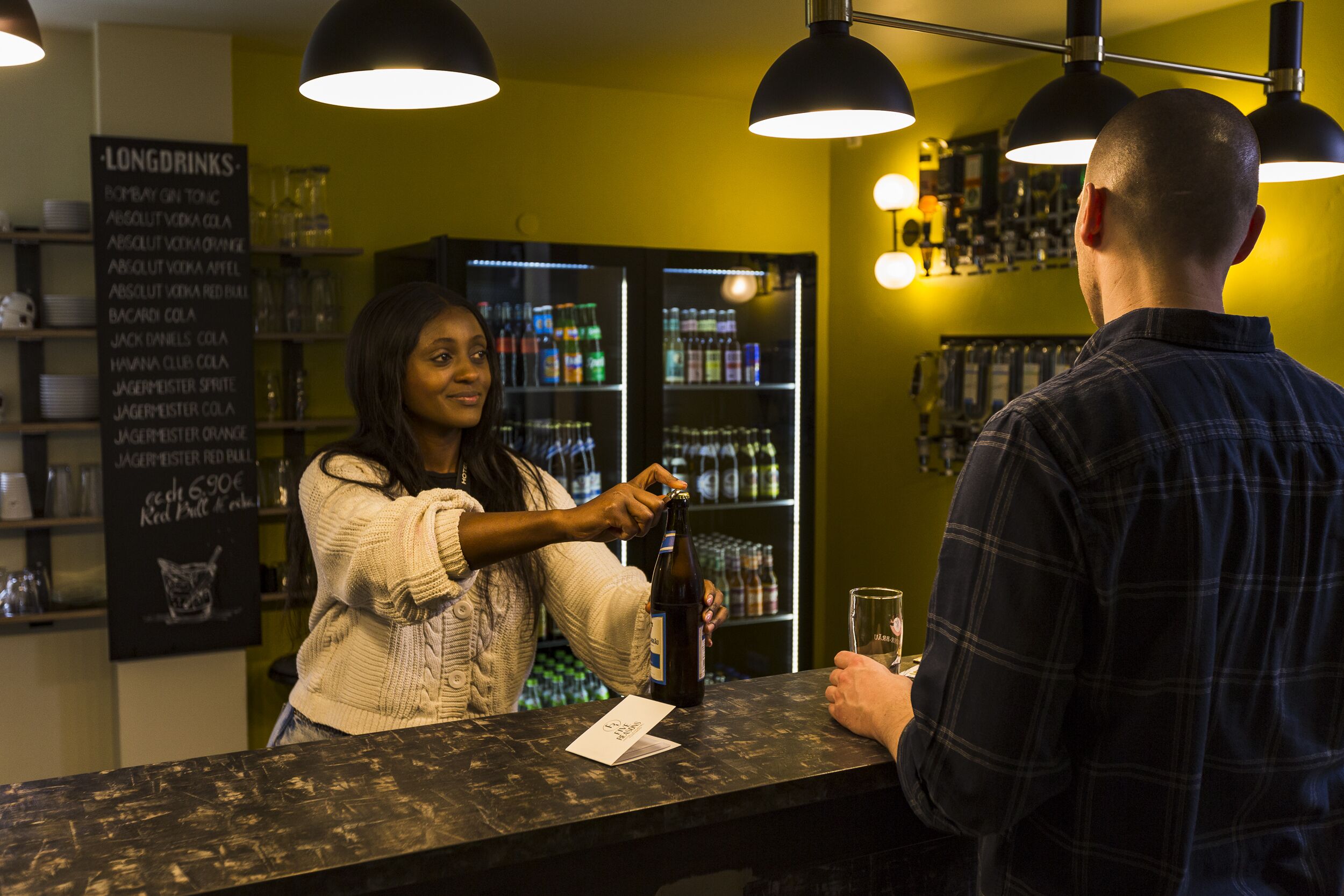 Zona de bar con un panel de menú que enumera bebidas, estantes con botellas y un refrigerador al fondo.
