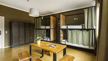 A dormitory room with bunk beds, a wooden table, and a bench. There are lockers and a hanging light fixture.