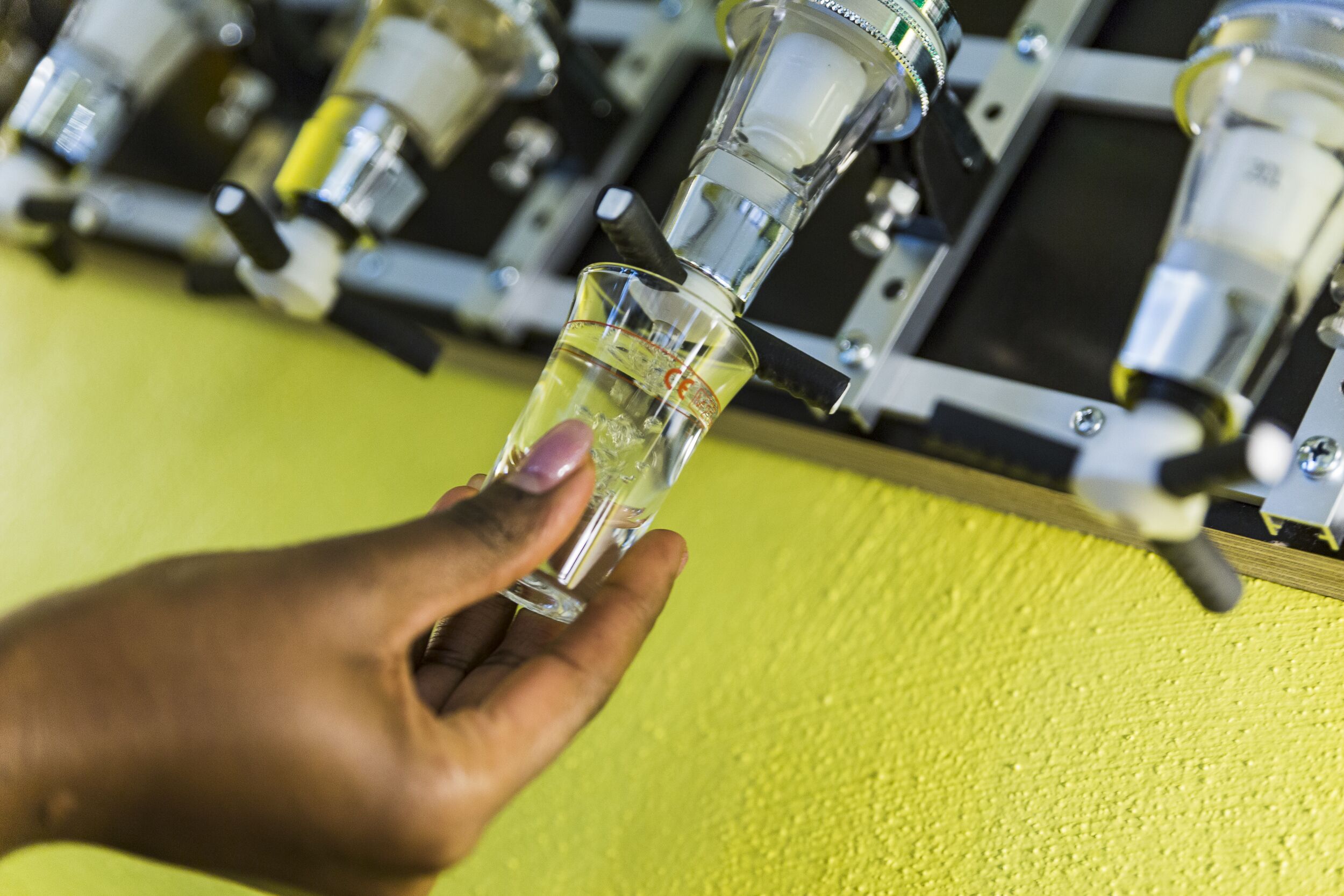 A hand holding a clear glass with a yellow liquid, possibly a drink, in front of a yellow wall with a metal structure behind it.