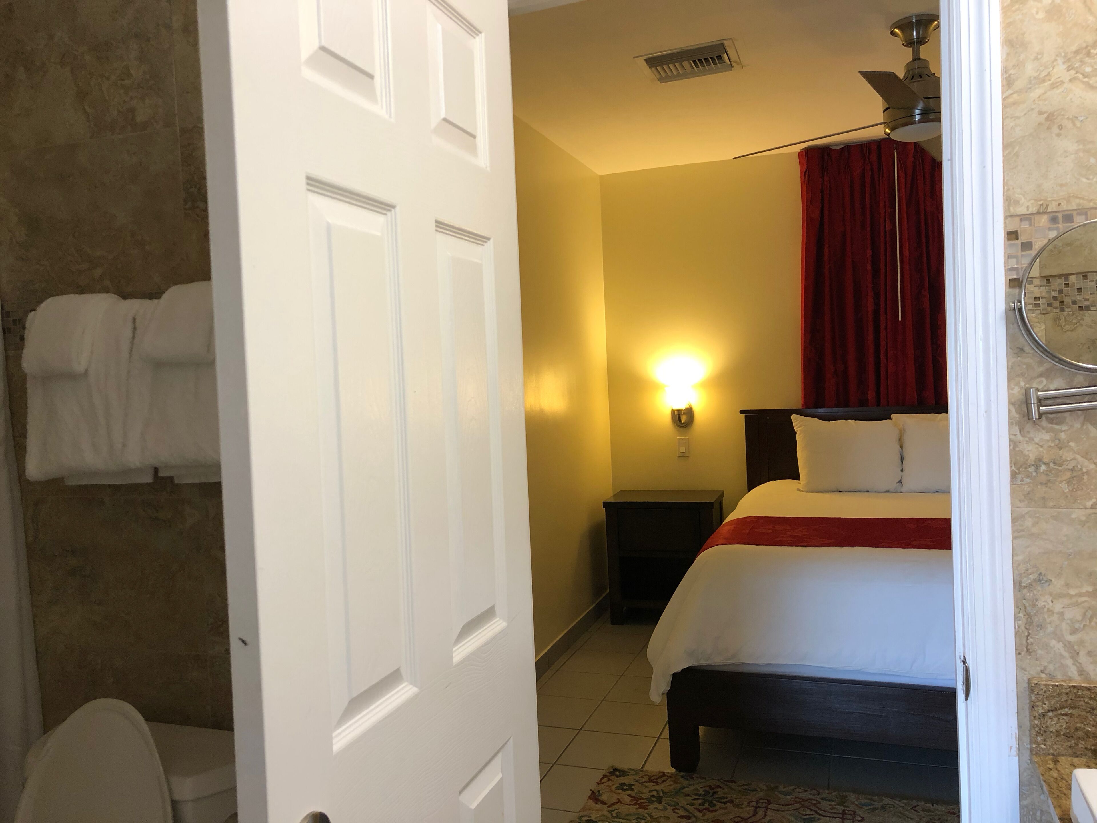 Une chambre d’hôtel avec un lit, une table de chevet, une lampe fixée au mur, un ventilateur de plafond et une salle de bain visible à travers une porte ouverte.