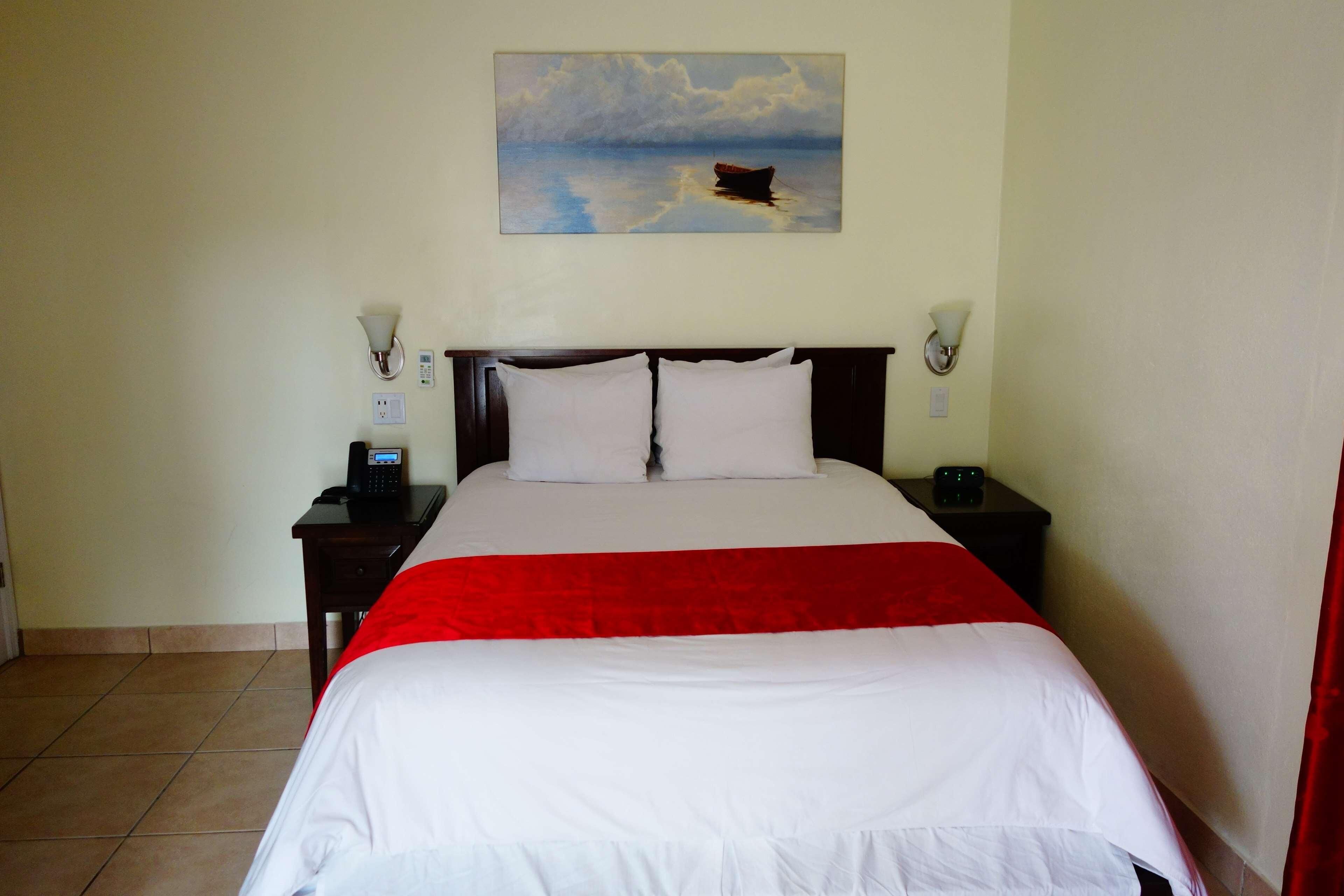 Habitación de hotel con una cama, dos mesitas de noche, un cuadro de un barco en el agua y una colcha roja y blanca.