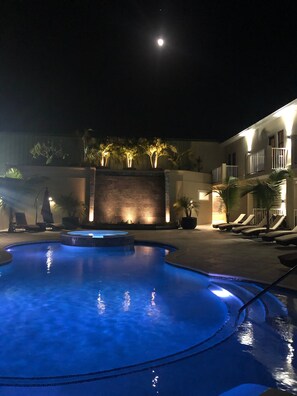 Un espace piscine éclairé pour la nuit, avec une lune dans le ciel, un bâtiment bien illuminé et des chaises longues.