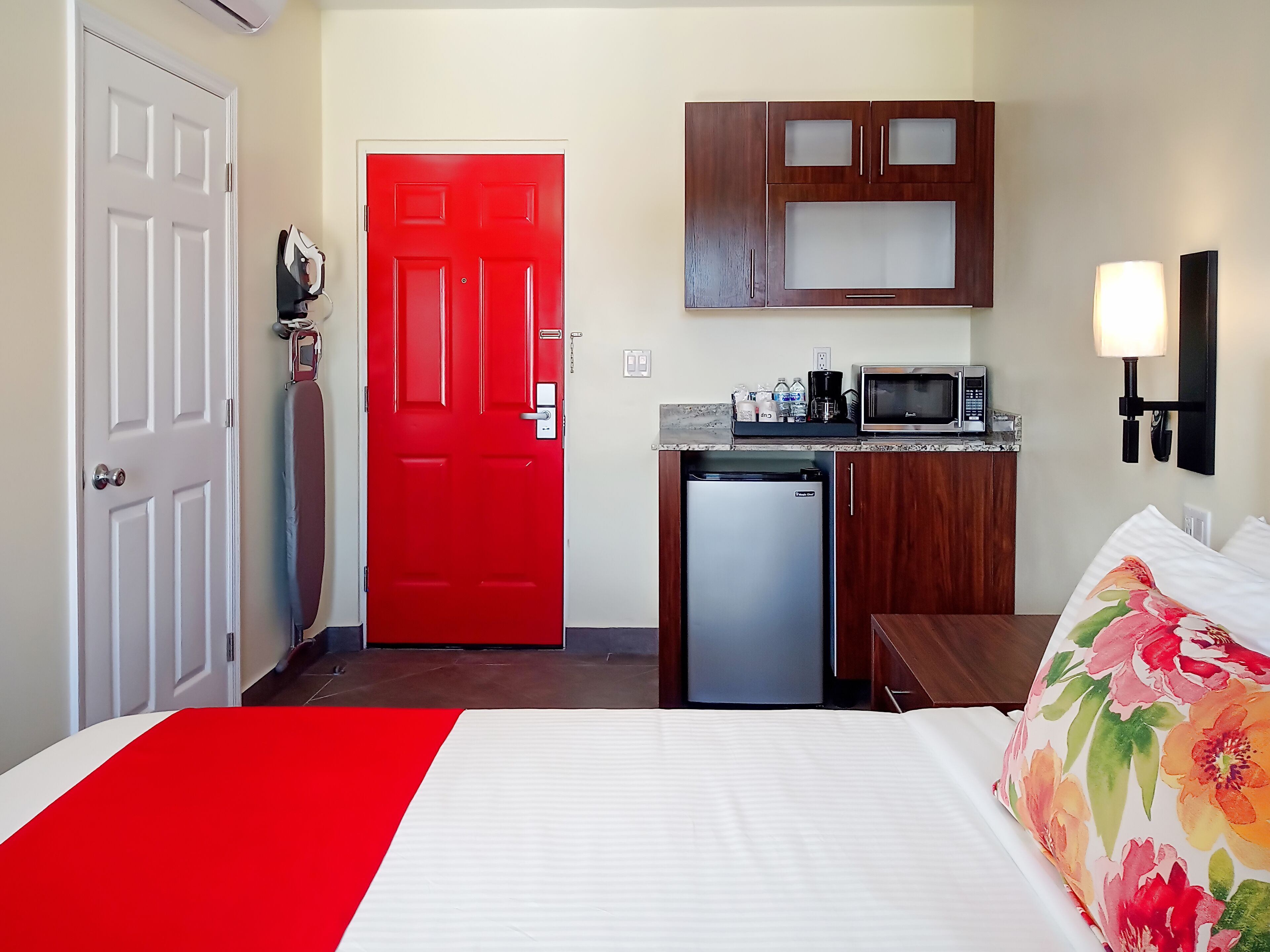 A compact hotel room with a red door, a small kitchenette, a bed with floral bedding, and a nightstand.