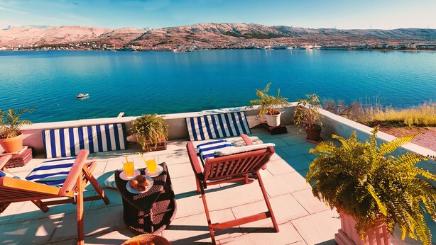 Terraço com cadeiras de descanso, uma mesa com bebidas e plantas em vasos, com vista para um lago e montanhas ao fundo.