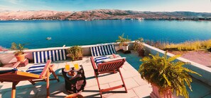 A terrace with lounge chairs, a table with drinks, and potted plants overlooking a lake with a mountainous backdrop. - Guest house Revelin (Pag)