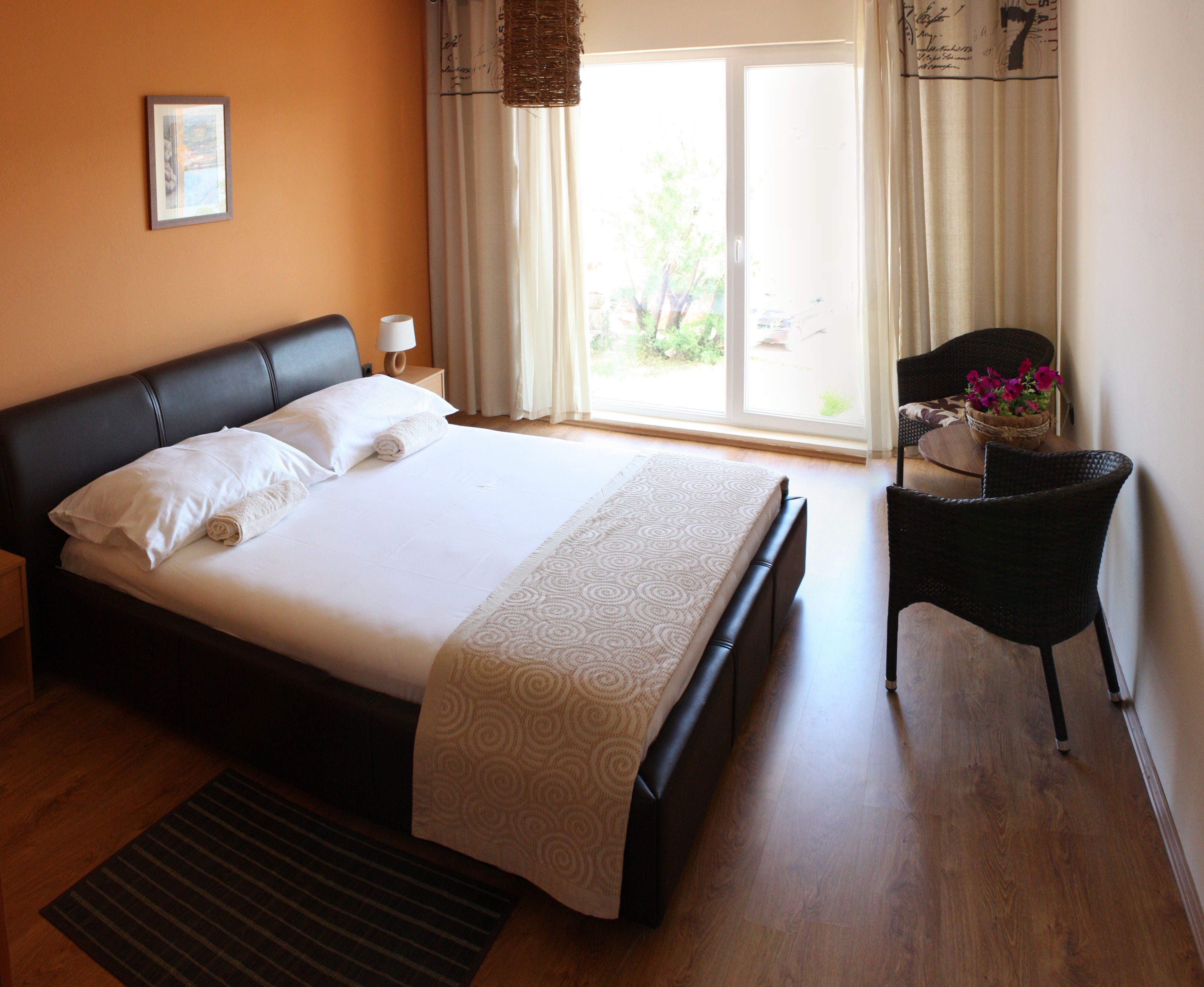 A neatly made bed with white linens, a dark headboard, and a patterned bedspread. There is a small side table with a lamp and a basket of flowers. The room has wooden flooring, a large window with curtains, and a view of greenery outside.