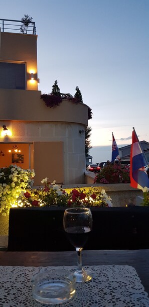 A table with a glass of water, a bench, and a view of a building with flags and flowers. - Guest house Revelin (Pag)