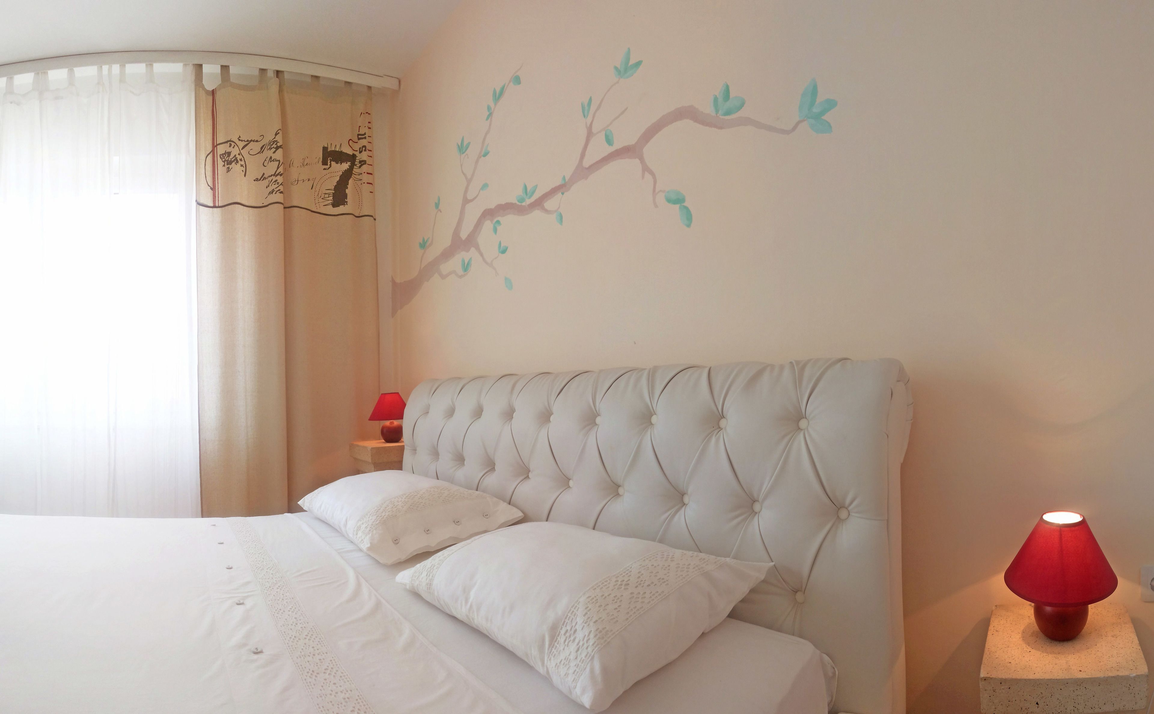 Ein Schlafzimmer mit einer gesteppten Kopfteil, weißer Bettwäsche, einer Wandsticker mit Zweigen, einer roten Lampe und einem Nachttisch mit einer roten Lampe.