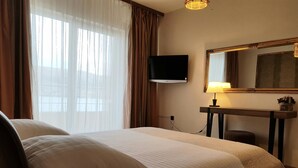 Quarto de hotel com uma cama, televisão fixada na parede, mesa de cabeceira com abajur e uma janela grande com cortinas.