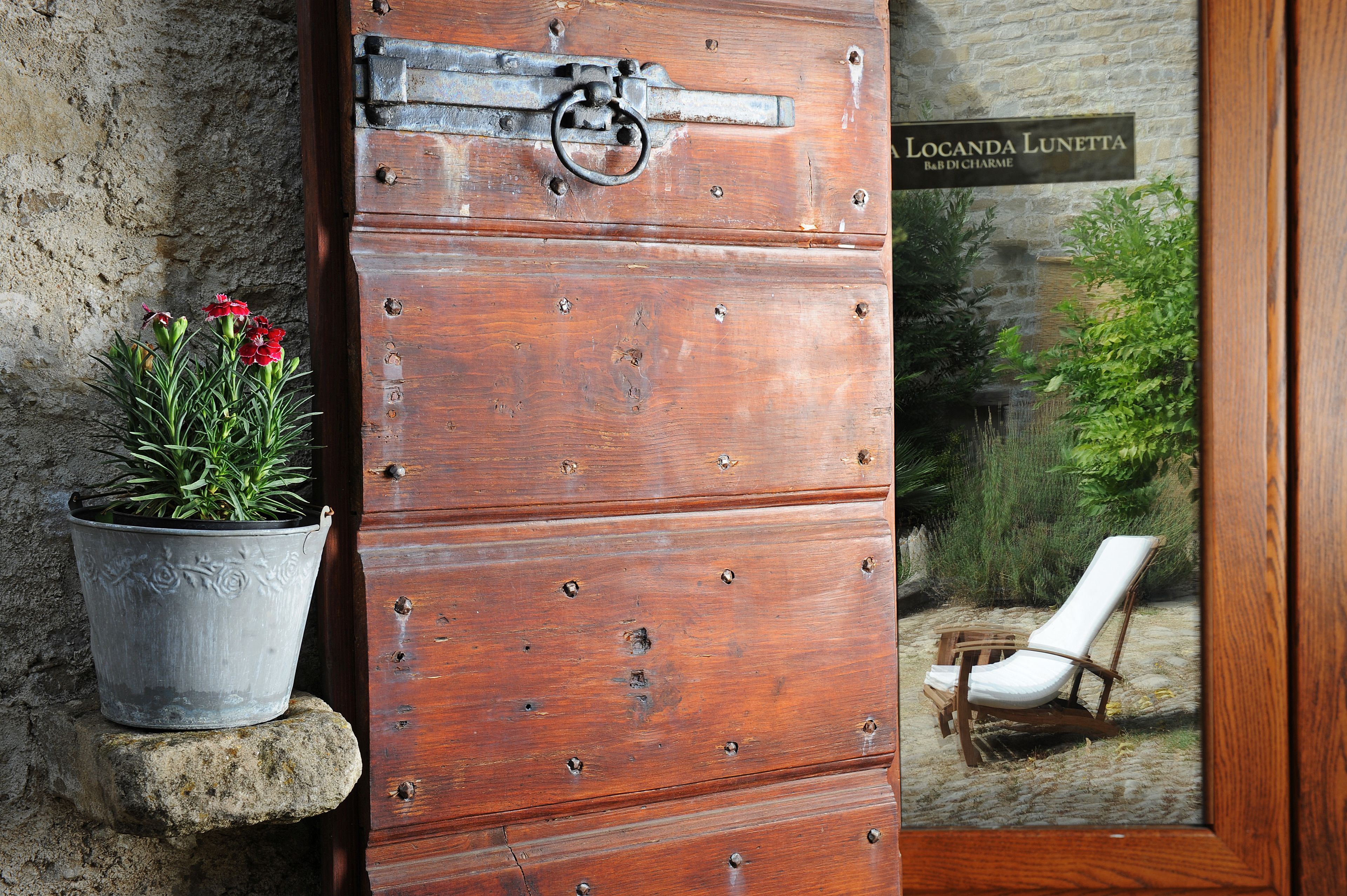 Una puerta de madera rústica con un pestillo metálico, una planta en maceta con flores rojas y un letrero que dice "Locanda Lunetta".