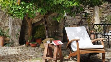 Ein Holzstuhl mit weißem Kissen, ein Korbwicker-Korb und ein Buch auf einem kleinen Holzschemel in einem Kopfsteinpflasterhof mit Topfpflanzen und einer Steinmauer.