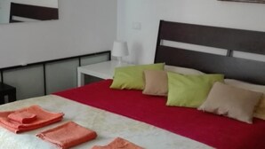 Una cama bien hecha con una colcha roja y varios almohadones, una mesita de noche con una lámpara y dos cuadros en la pared.