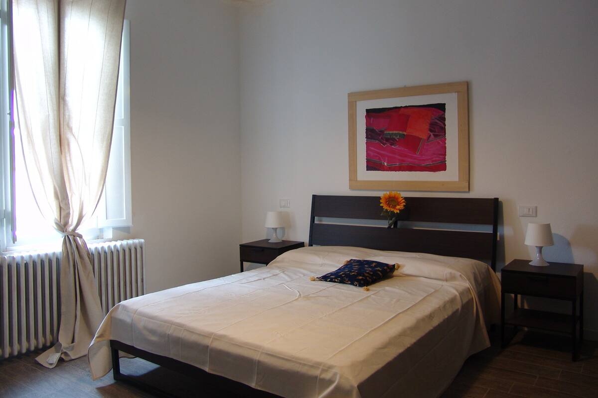 A neatly made bed with a single pillow, a framed picture on the wall, and a radiator beside the window.