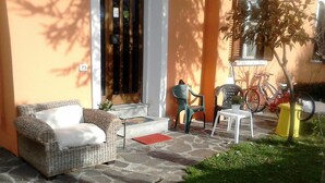 Un patio empedrado con un sofá de mimbre y dos sillones, una mesita y una bicicleta estacionada frente a un edificio con puerta amarilla.