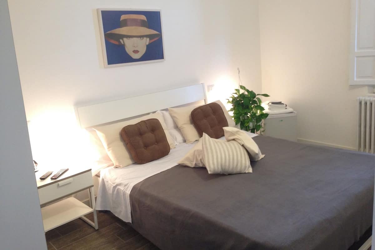 A neatly made bed with a grey bedspread and brown headboard pillows, a bedside table with a lamp, and a framed picture on the wall.