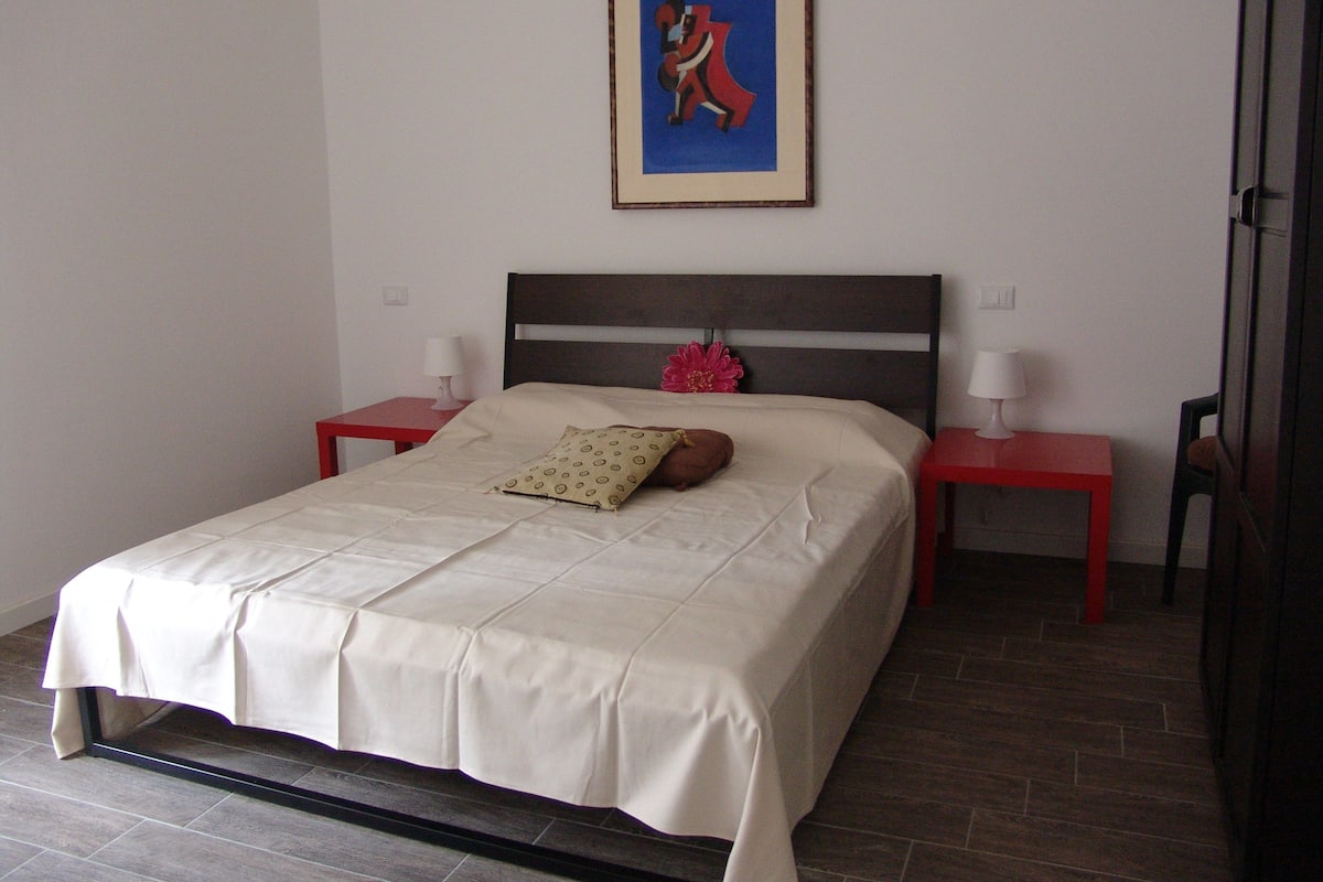 A neatly made bed with a beige bedspread and two pillows, flanked by two red nightstands with lamps, in a room with a simple, clean design.