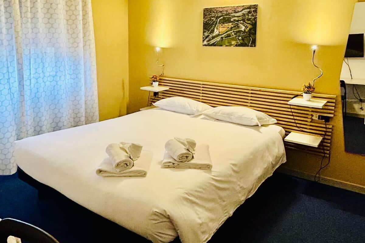 A hotel room with a bed, two bedside tables, a desk, a chair, and an air conditioning unit.