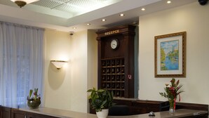 A reception area with a reception desk, a clock, a wall-mounted light, a framed painting, and a potted plant. - Villa Lauretta (Karlovy Vary)