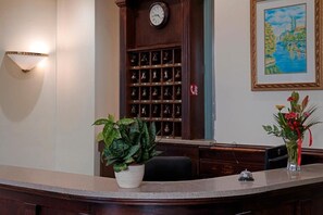 A reception desk with a clock, a wall-mounted light, a plant, a vase with flowers, and a framed painting. - Villa Lauretta (Karlovy Vary)