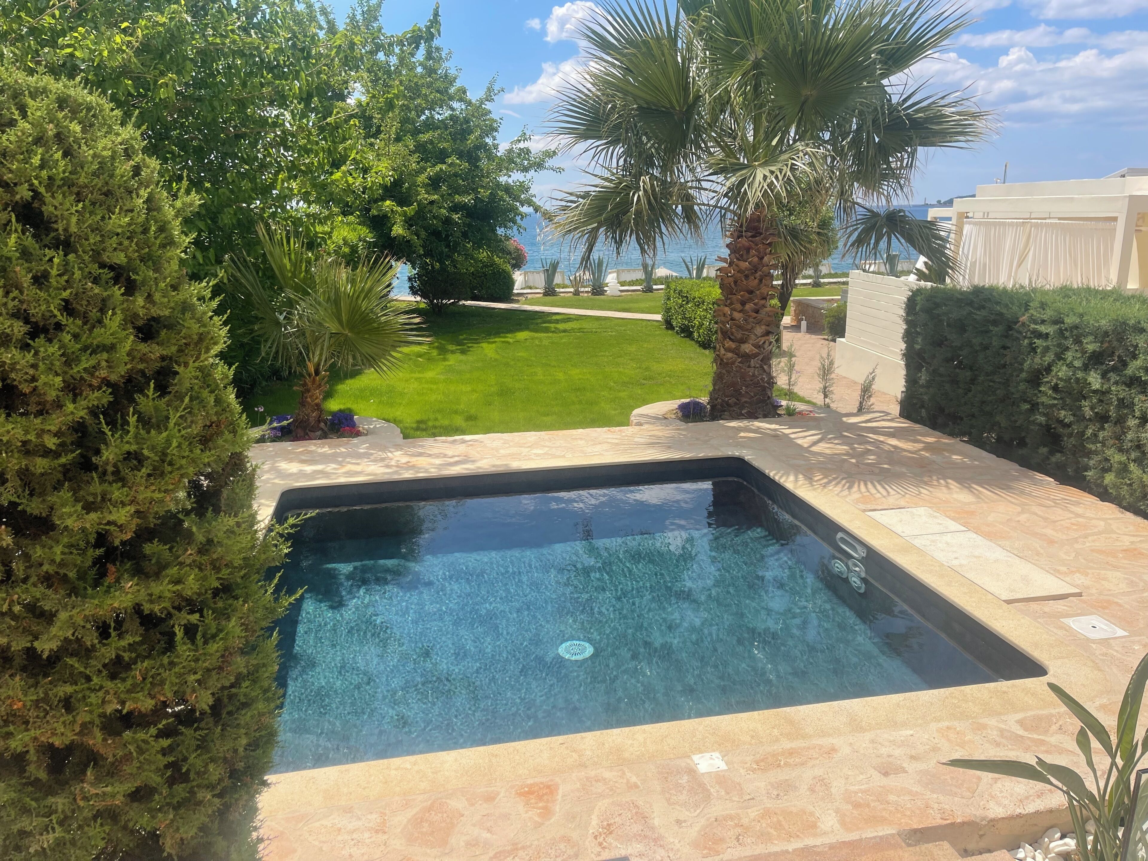 Una piscina con vistas despejadas al océano, rodeada de vegetación cuidada y una zona de patio empedrada.