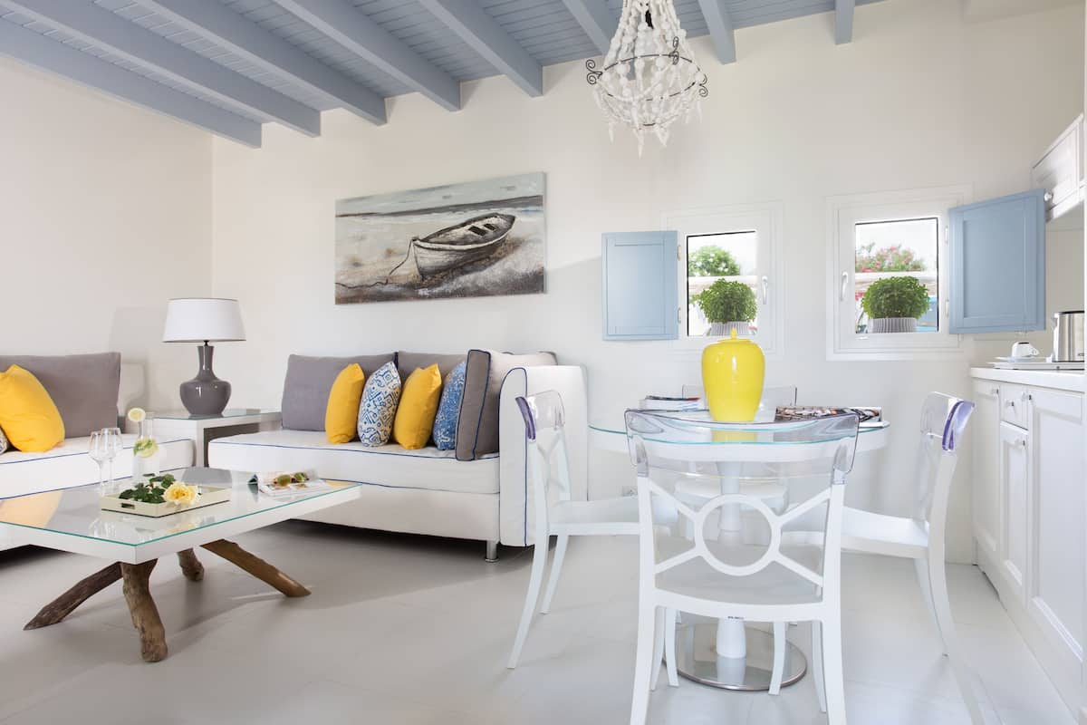 Un salon moderne avec une table basse en verre, des canapés blancs ornés de coussins jaunes, un coin repas avec une table et des chaises en verre, et une cuisine avec des meubles blancs et des volets bleus.