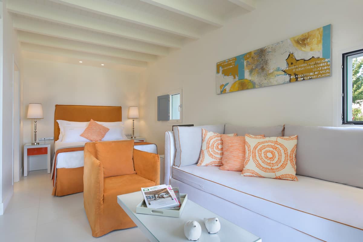 Une chambre d’hôtel moderne, équipée d’un lit, d’un canapé, d’un fauteuil et d’une table basse. Un tableau orne le mur et une fenêtre laisse entrer la lumière naturelle.