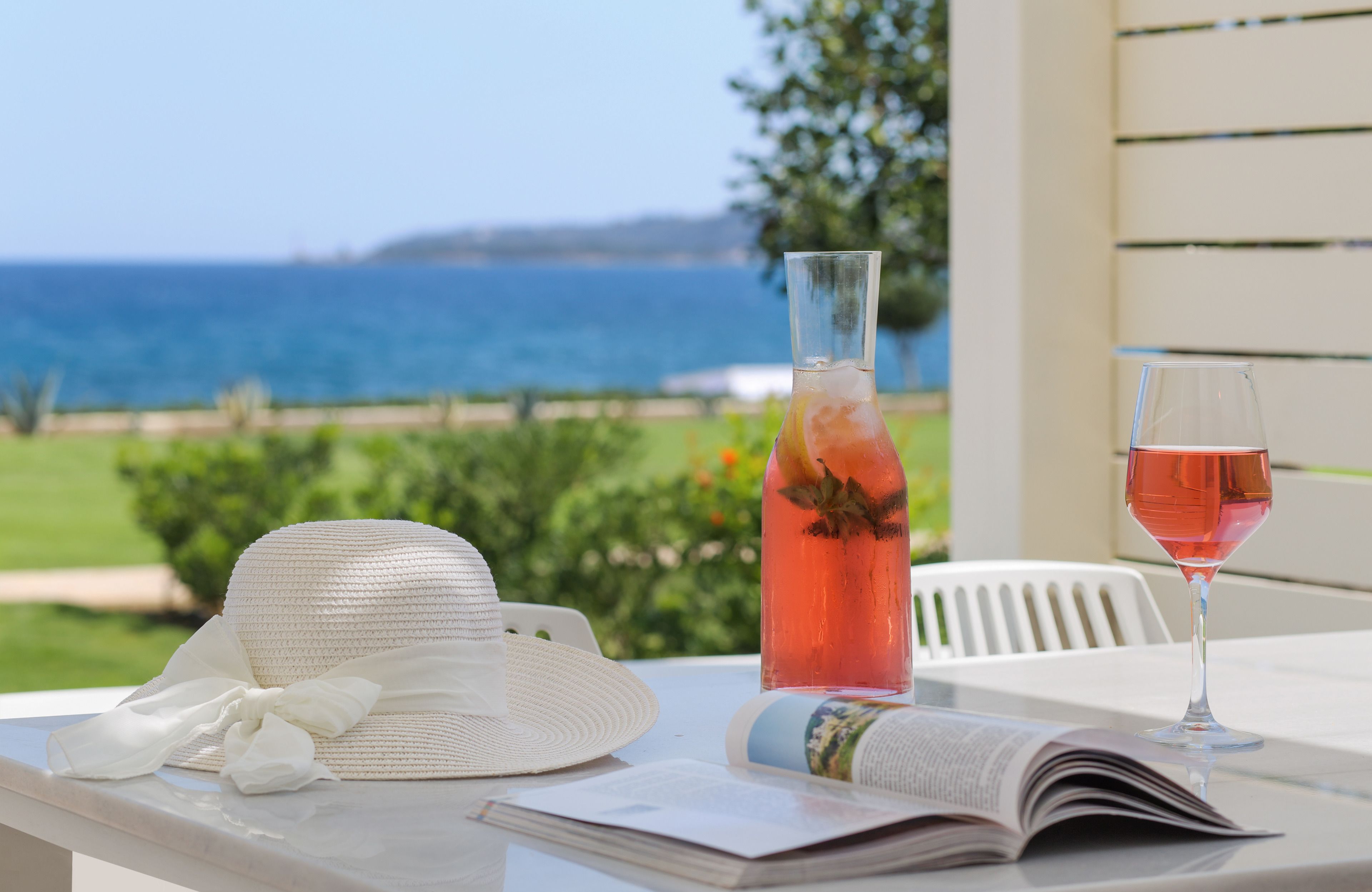 A table with a white hat, a bottle of pink drink, and a glass of pink drink, with a view of the ocean in the background.