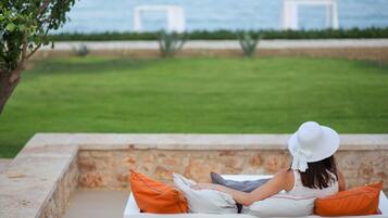 A person sitting on a white bench with orange cushions, overlooking a grassy area with a stone wall and a view of the sea.