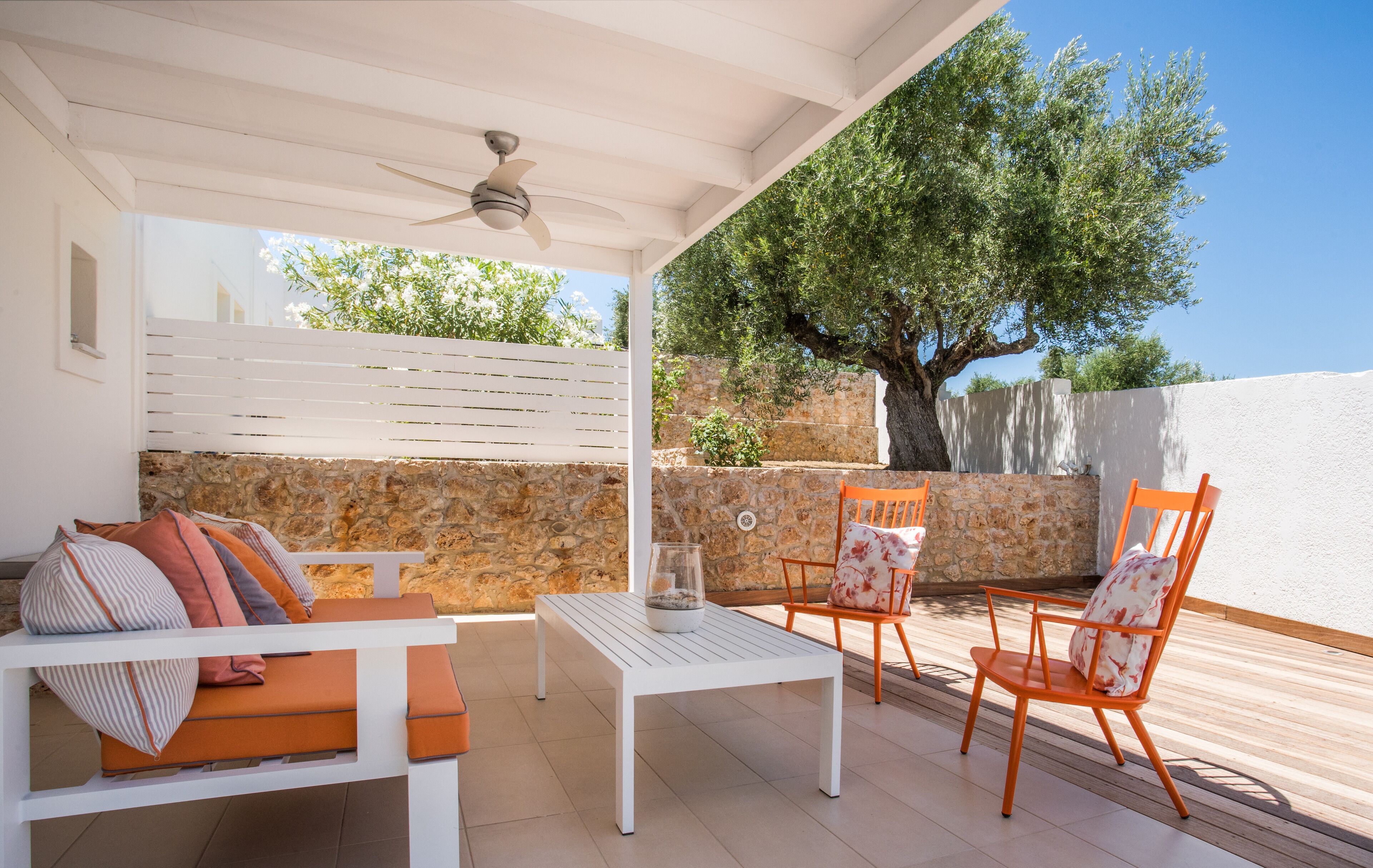 Un espace extérieur abrité avec des tables et des chaises, la table étant blanche et les chaises orange, un ventilateur de plafond et un mur en pierre, avec un arbre en arrière-plan.