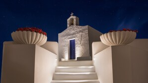 A white building with a blue door and a cross on top, surrounded by white walls and two large flower pots.