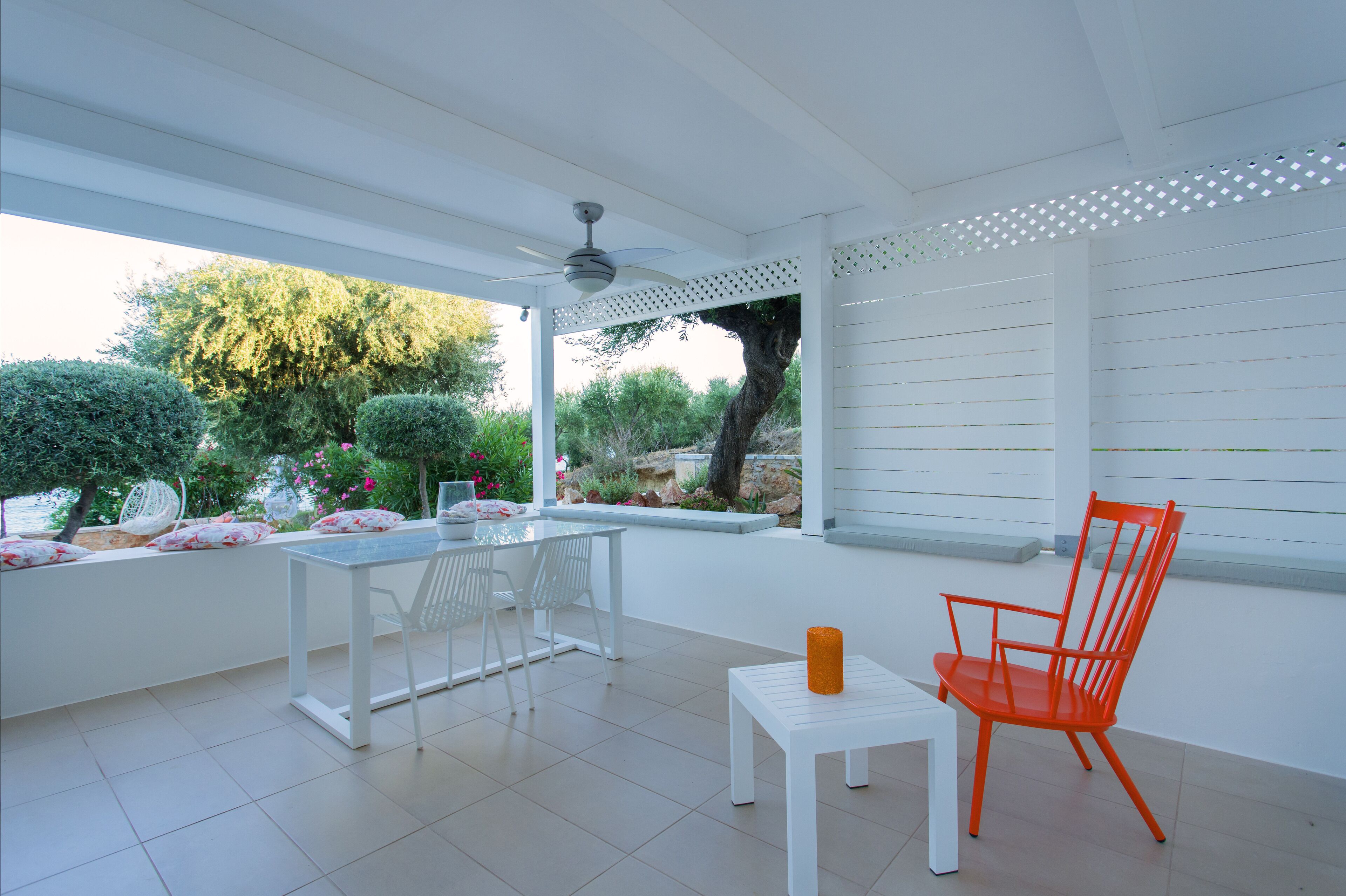 Un patio techado con una mesa y sillas blancas, una silla naranja brillante y una pequeña mesa con una vela.