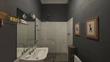 A modern bathroom with a white sink, mirror, and shower. There are framed pictures on the wall and a towel rack with bottles.