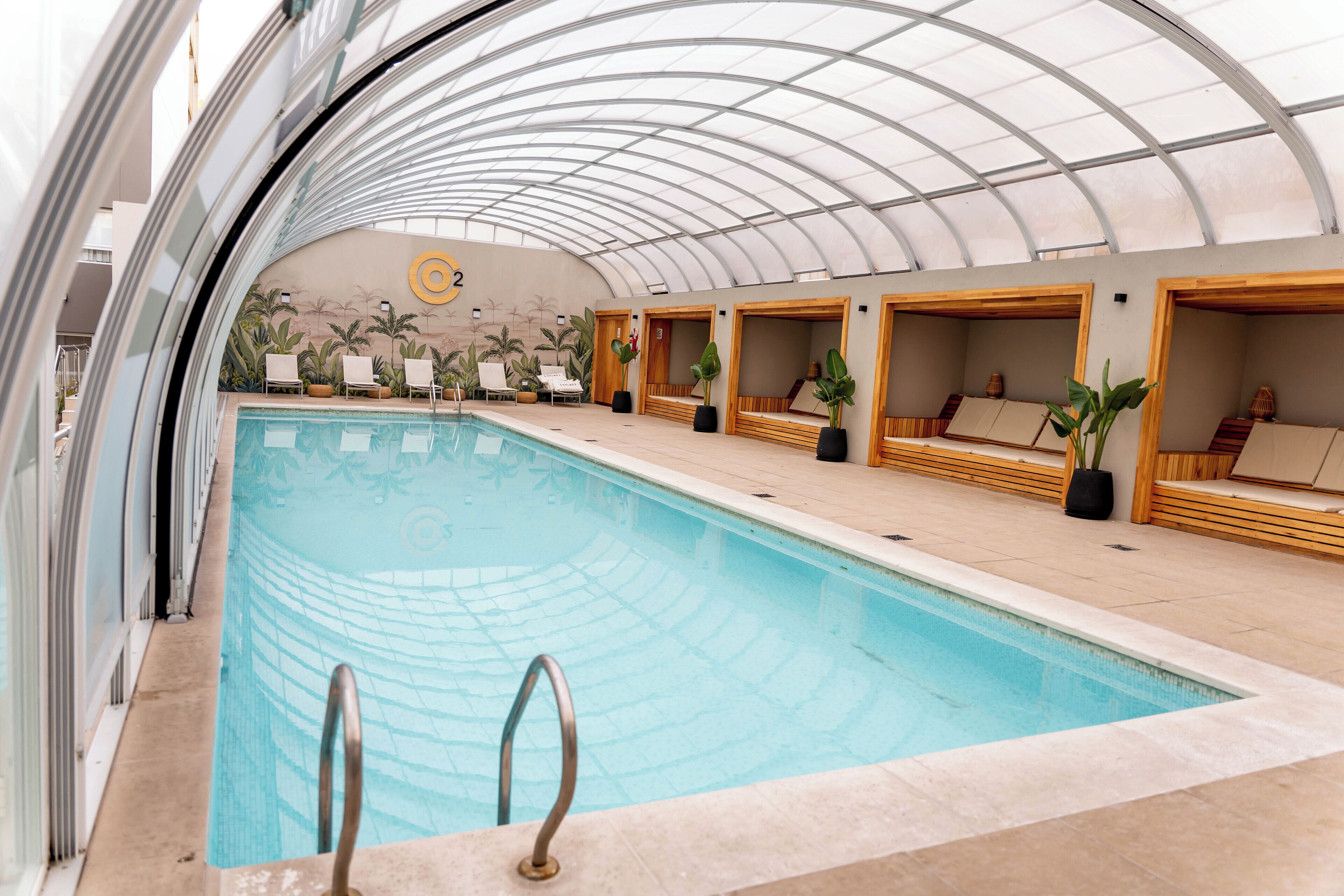An indoor swimming pool with a glass ceiling, lounge chairs, and wooden seating areas.