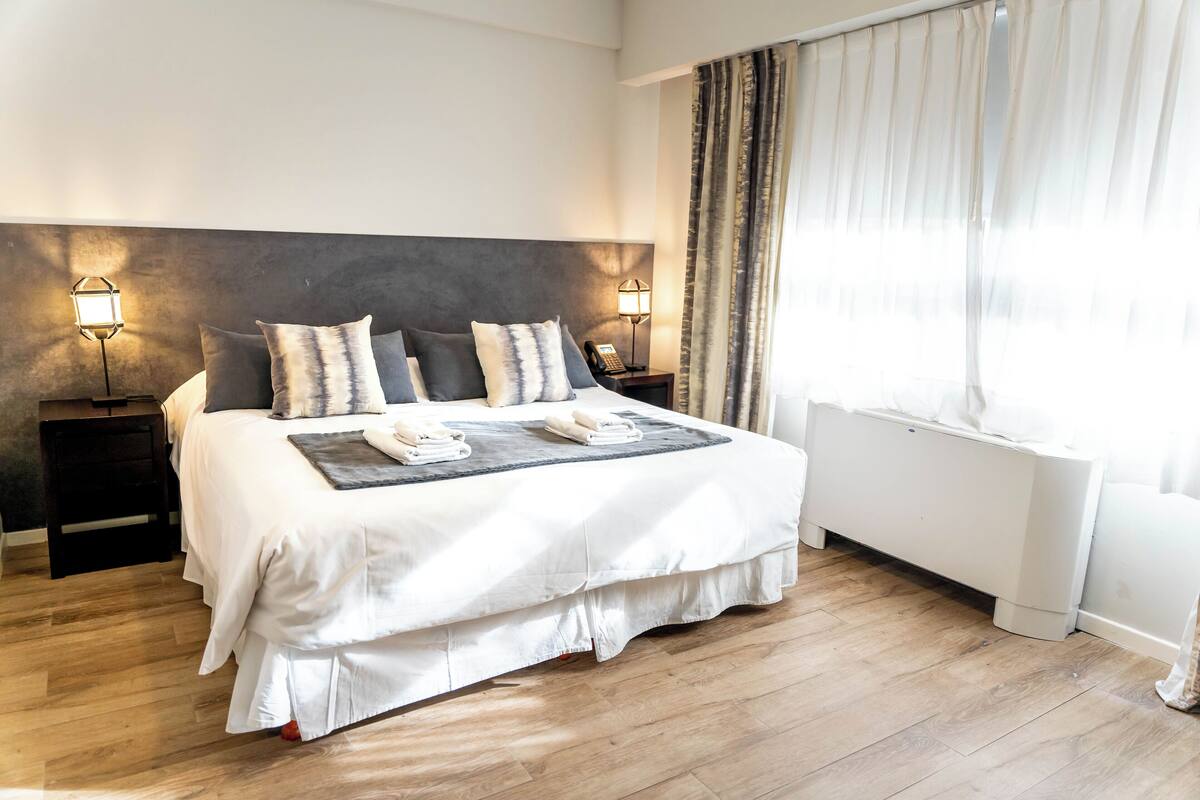 A neatly made bed with a grey headboard, two bedside tables with lamps, a window with sheer curtains, and a radiator.