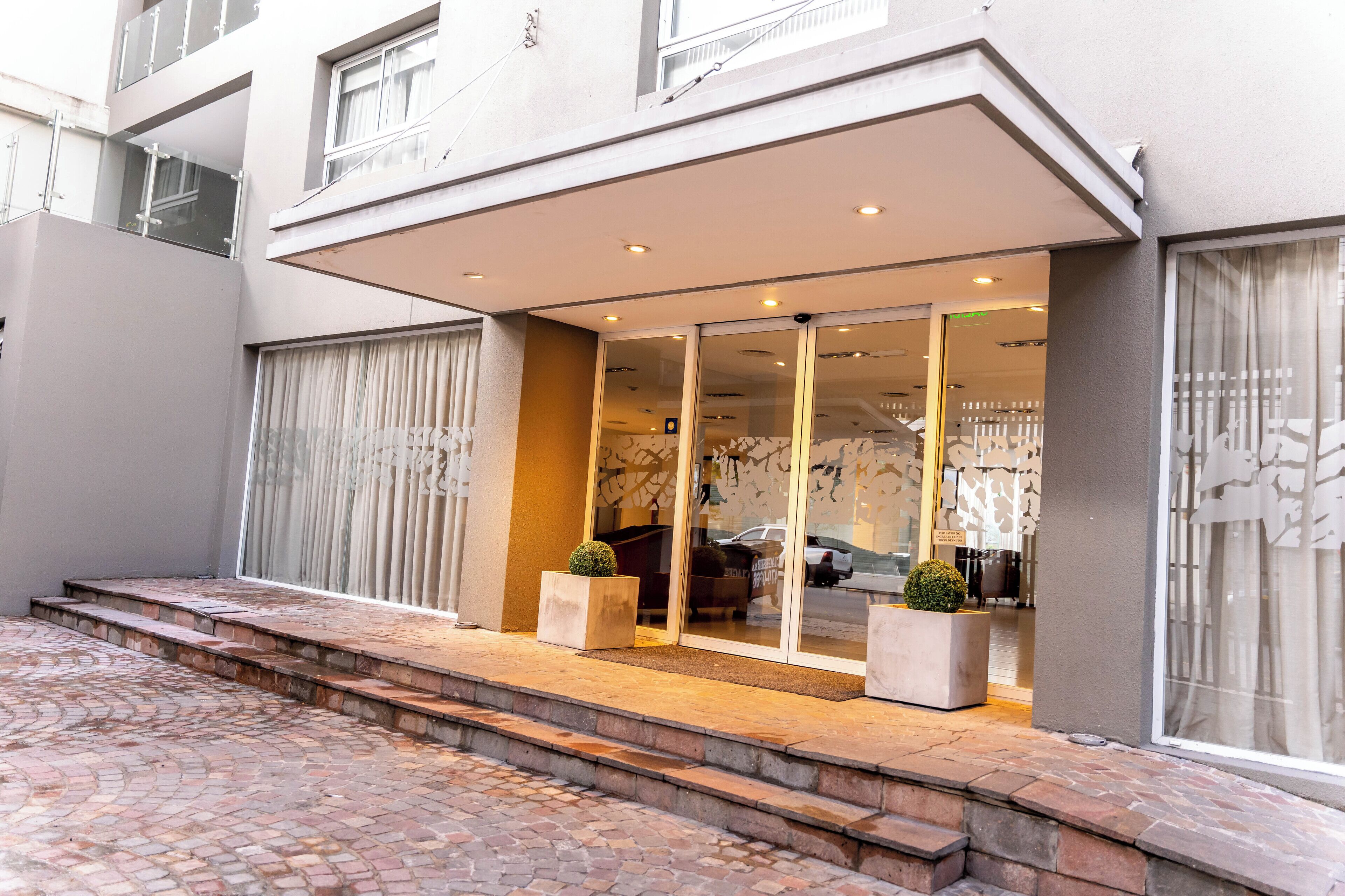 A modern building with a glass entrance, steps leading up to the door, and a small garden area with potted plants.