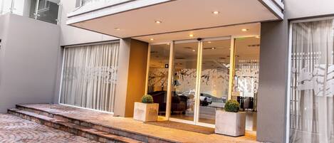 A modern building with a glass entrance, steps leading up to the door, and a small garden area with potted plants.