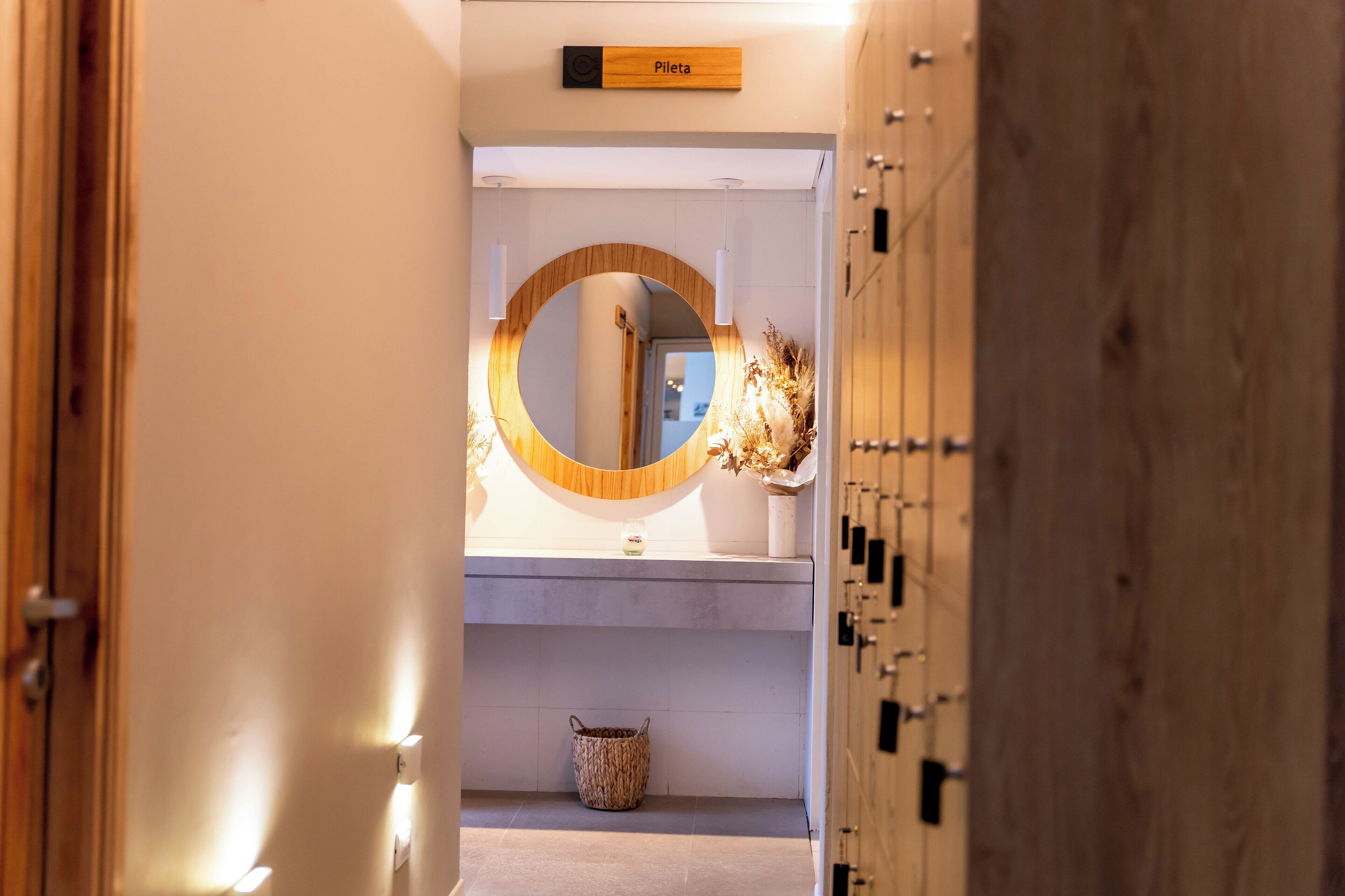 A hallway with a wooden door labeled "Piletas," a round mirror, a small woven basket, and a wall-mounted light fixture.