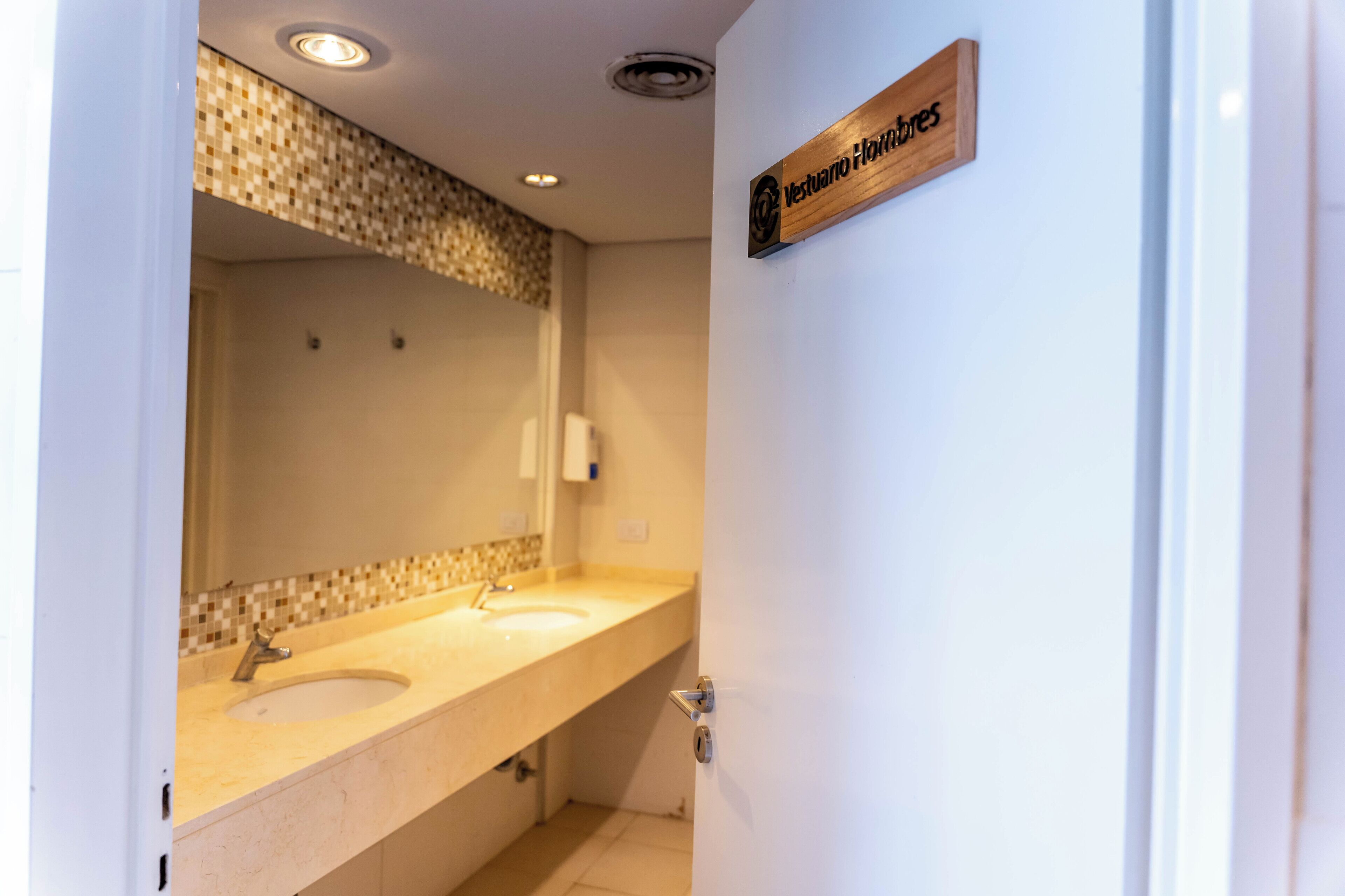 A bathroom with a double sink, a mirror, and a wooden sign above the door.